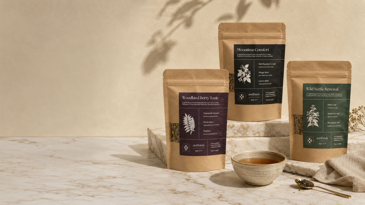 Three stand-up pouches of herbal tea in kraft paper packaging are arranged on a marble surface with a small bowl of tea and a spoon. The pouches have dark labels with botanical illustrations and text including 'Woodland Herb Tea', 'Mountain Comfort', and 'Wild Sage Herbal'.