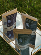 Three packages of 'Woodland Berry Tonic', 'Moontime Comfort', and 'Wild Nettle Renewal' on a reflective surface with grass in the background.